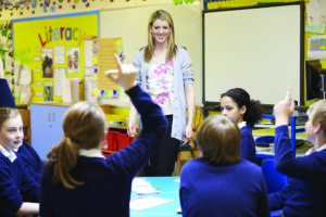 Classroom with Teacher Standing and Children Sitting with their Hands Up